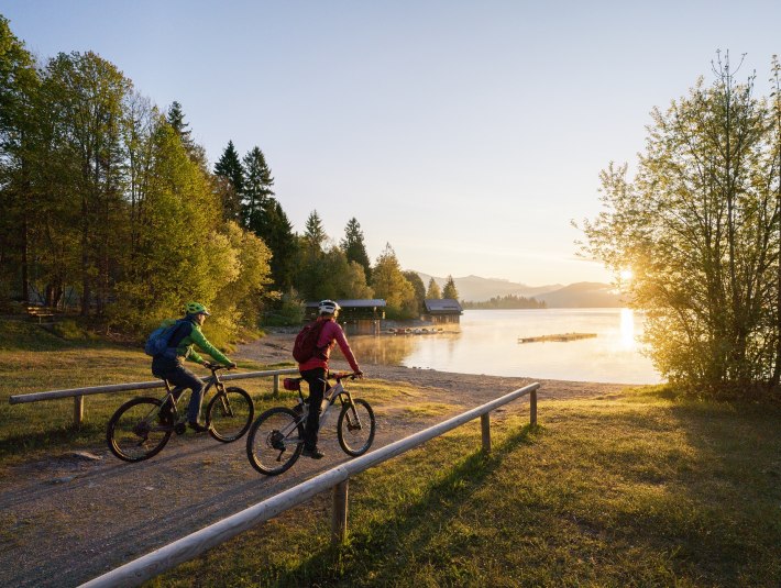 Welch Ausblick auf den Walchensee! Zwei Fahrradfahrer fahren auf den in morgendlichen Sonnenlicht getauchten Walchensee zu., © Archiv Tölzer Land Tourismus| Foto: Bernd Ritschel