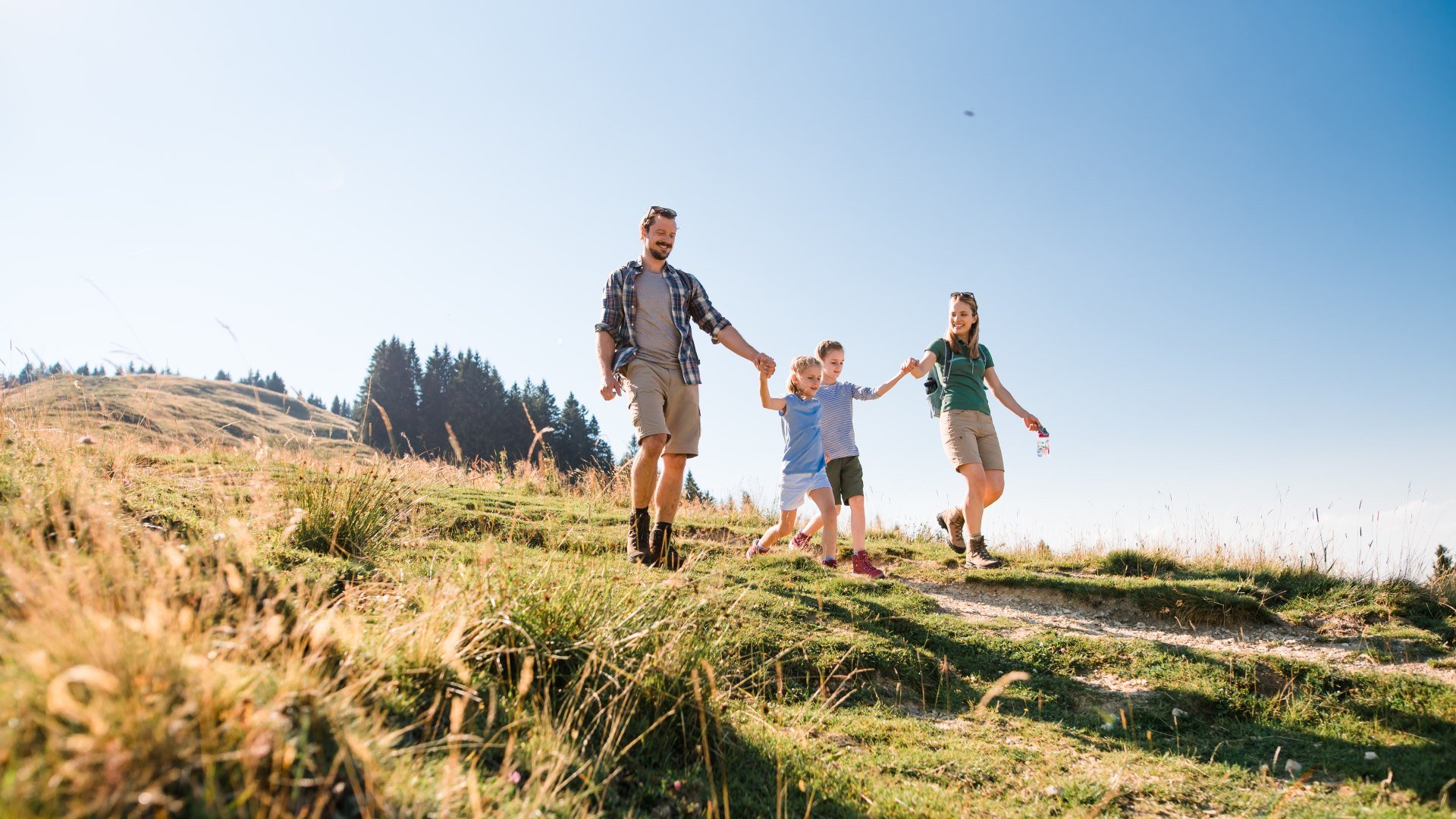 Eine vierköpfige Familie wandert Hand in Hand auf dem Wanderweg einer Almwiese, © Tölzer Land Tourismus Foto: Leonie Lorenz