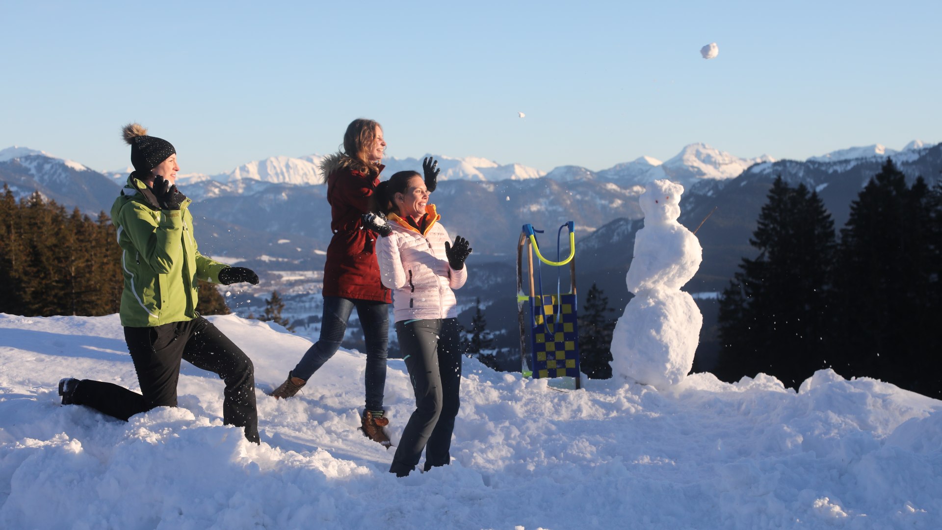 Das Bild zeigt drei Frauen in Winterkleidung, die Schnebälle in Richtung eines Rodels und Schneemannes werfen. Im Hintergrund sieht man die winterliche Bergkulisse, © Tourist Information Bad Tölz Foto: Hias Krinner